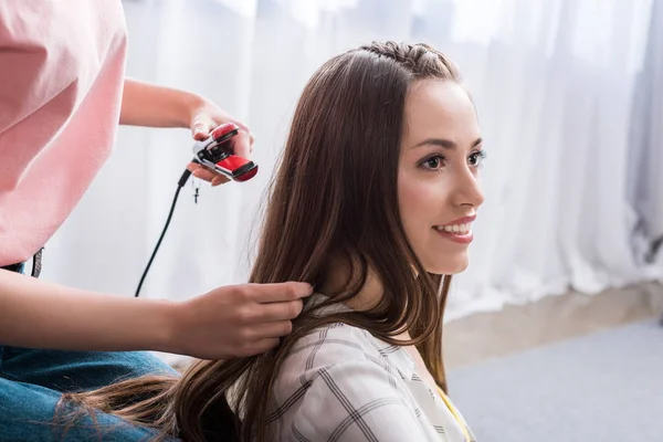 Обрезанный снимок женщины парикмахерской подруги с помощью выпрямителя для волос на диване дома — стоковое фото