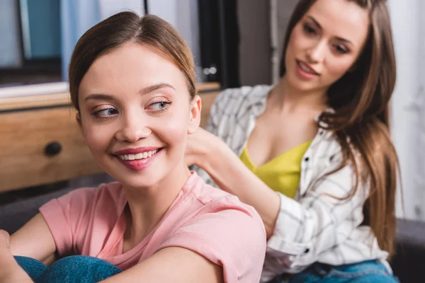 Улыбающаяся женщина парикмахерша подруги по волосы выпрямитель на диване дома — стоковое фото