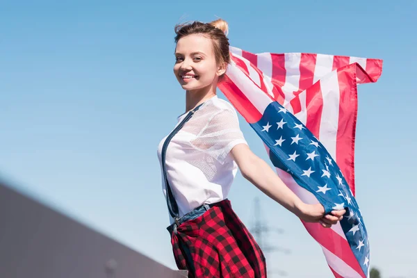 Sonriente joven mujer sosteniendo bandera americana contra el cielo azul, concepto del día de la independencia - foto de stock