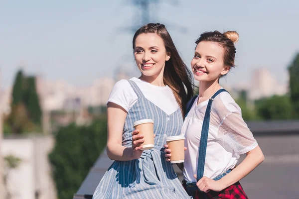 Улыбающиеся юные подружки с бумажными чашками кофе на крыше — стоковое фото