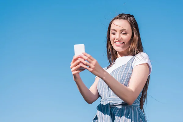 Atractiva joven mujer tomando selfie en el teléfono inteligente contra el cielo azul - foto de stock