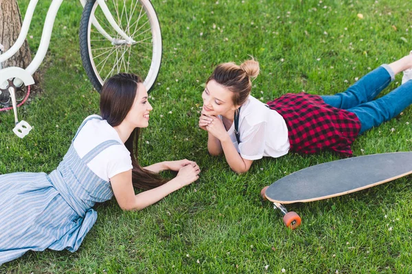 Vista elevada de sonrientes amigas tendidas en la hierba con bicicleta y monopatín en el parque - foto de stock