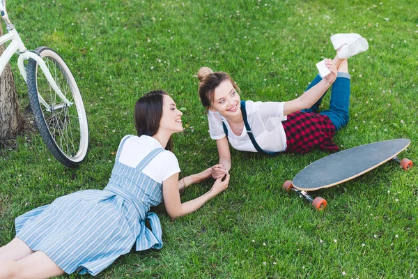 Vue grand angle de la femme souriante prenant selfie sur smartphone avec amie sur herbe avec planche à roulettes et vélo dans le parc — Photo de stock