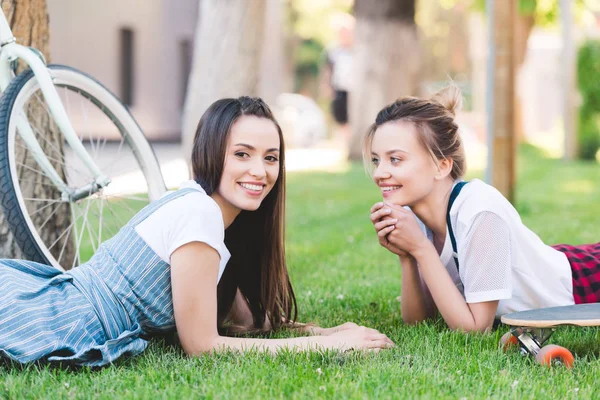 Amiche sorridenti sdraiate sull'erba con bicicletta e skateboard nel parco — Foto stock