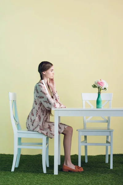 Stylisches Teenie-Mädchen mit Zöpfen sitzt am Tisch mit Blumen, auf gelb — Stockfoto