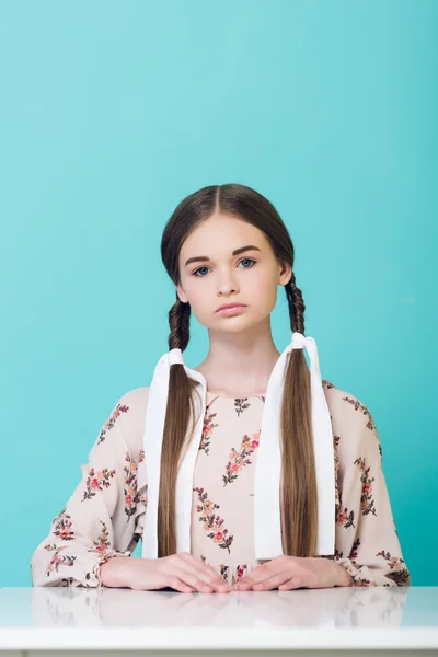 Schönes Teenie-Mädchen mit Zöpfen sitzt am Tisch, isoliert auf Türkis — Stockfoto