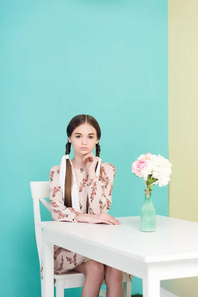 Attraktive Teenie-Mädchen mit Zöpfen im Sommerkleid sitzt am Tisch mit Blumen, auf blau — Stockfoto