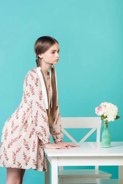 Adolescente chica con trenzas de pie en la mesa con ramo de flores, aislado en azul - foto de stock