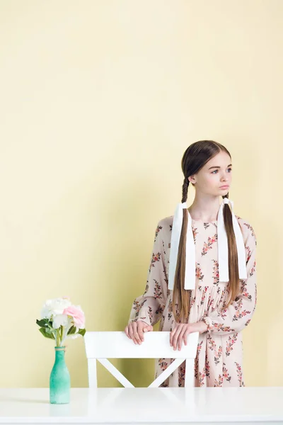 Schöne Teenie-Mädchen mit Zöpfen posiert an Stuhl und Tisch mit Blumen, auf gelb — Stockfoto