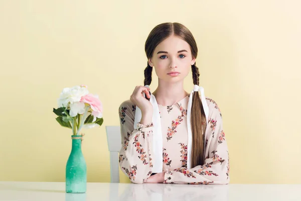 Elegante ragazza adolescente con trecce seduto a tavola con vaso e fiori, isolato su giallo — Foto stock