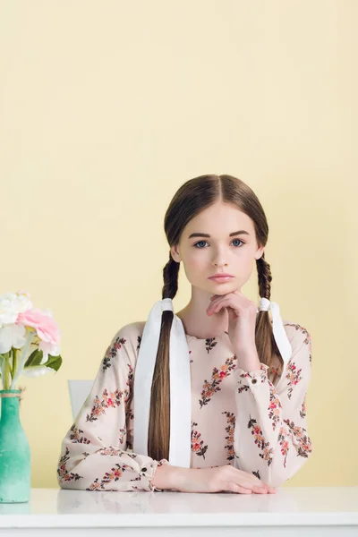 Schöne junge Mädchen mit Zöpfen sitzen am Tisch mit Blumen, isoliert auf gelb — Stockfoto