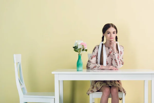 Fashionable teen girl with braids sitting at table with flowers, on yellow — Stock Photo
