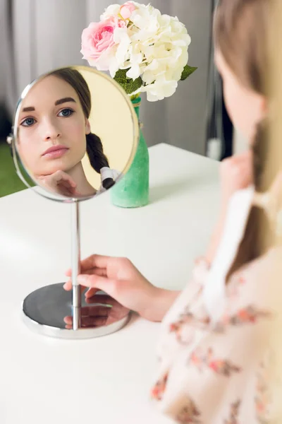 Селективное внимание девушки-подростка с косичками, сидящей за столом и смотрящей в зеркало — стоковое фото