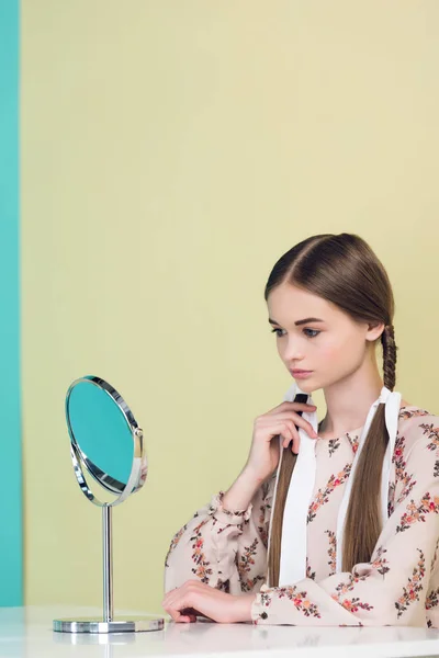 Teen girl guardando specchio e toccando le trecce, su giallo — Foto stock