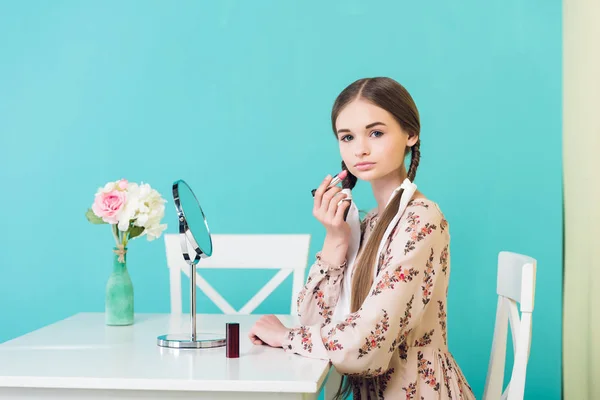 Attractive stylish teen girl applying lipstick with mirror, on blue — Stock Photo