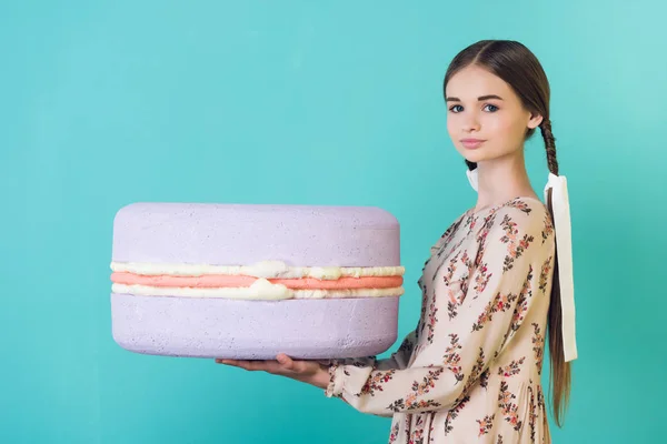 Belle jeune fille élégante avec des tresses tenant grand macaron violet, isolé sur turquoise — Photo de stock