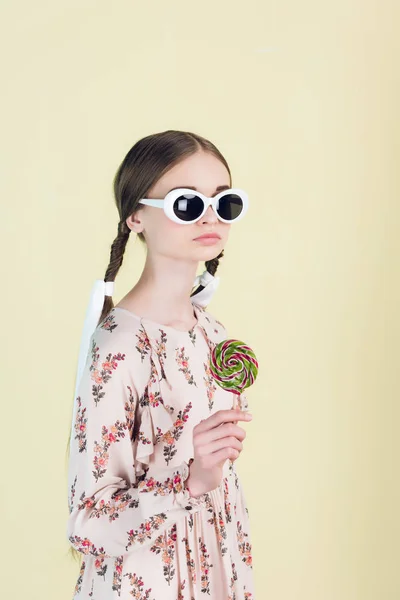 Fashionable youth girl in sunglasses with braids holding lollipop, isolated on yellow — Stock Photo