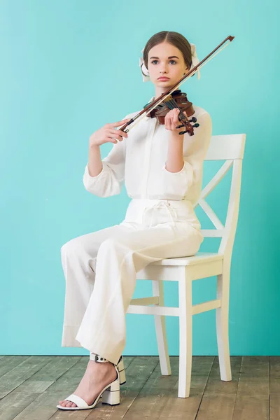 Attractive teen musician playing violin and sitting on chair, on blue — Stock Photo