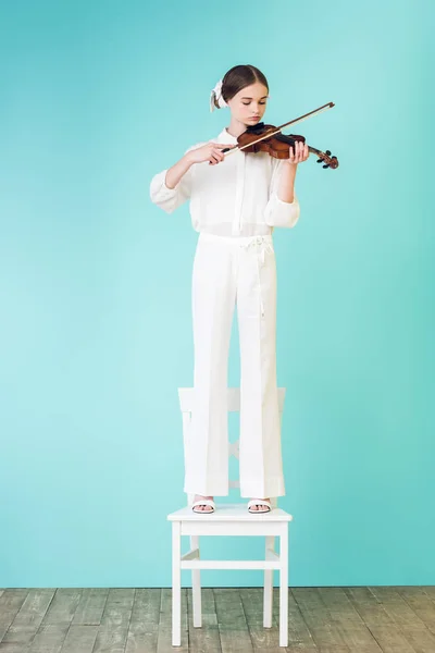 Teenager Mädchen in weißem Outfit spielt Geige und steht auf Stuhl, auf Türkis — Stockfoto