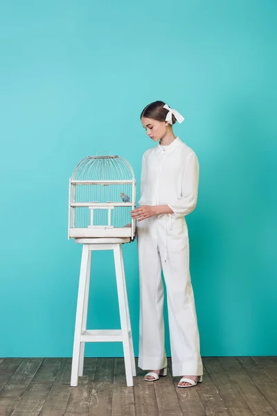 Jeune fille à la mode en tenue blanche élégante regardant perroquet en cage, sur turquoise — Photo de stock