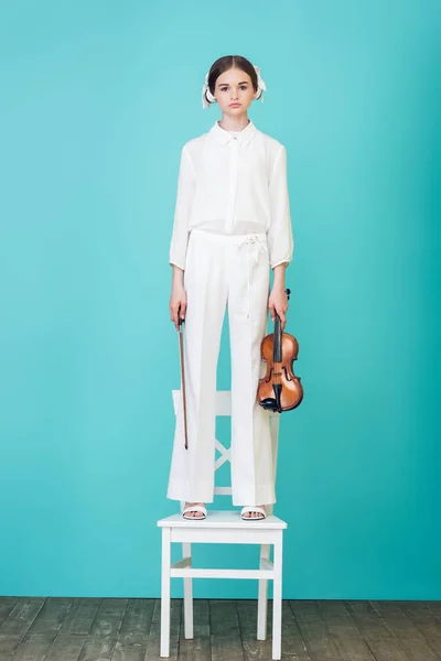 Mädchen im weißen Outfit, Geige haltend und auf Stuhl stehend, auf blau — Stockfoto