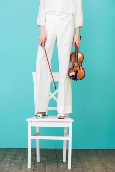 Cropped view of girl holding violin and standing on chair, on blue — Stock Photo