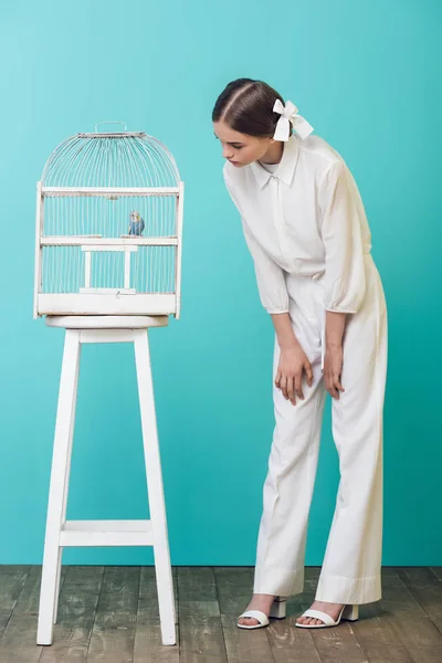 Привлекательная девушка в белом смотрит на попугая в клетке, на бирюзовый — стоковое фото