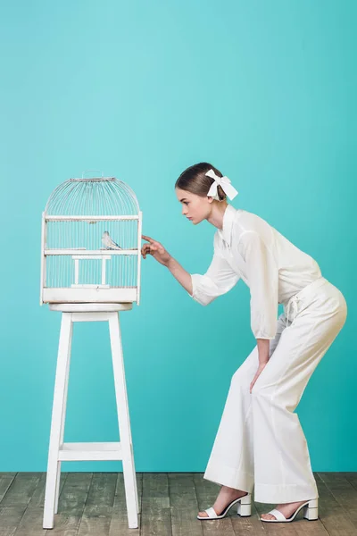 Belle fille en tenue blanche regardant perroquet en cage, sur turquoise — Photo de stock