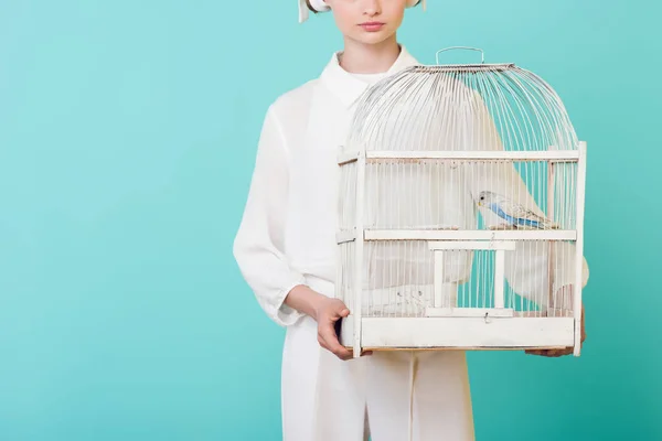 Обрезанный вид девочки в белой клетке с попугаем, изолированной на бирюзе — стоковое фото