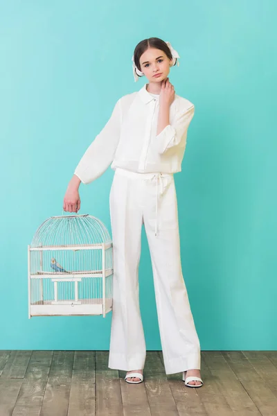 Beautiful girl in white outfit with parrot in cage, on turquoise — Stock Photo