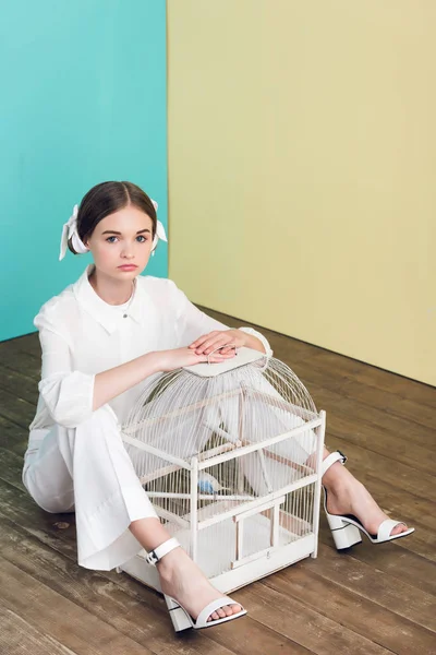 Stylish teen girl with parrot in cage sitting on floor — Stock Photo