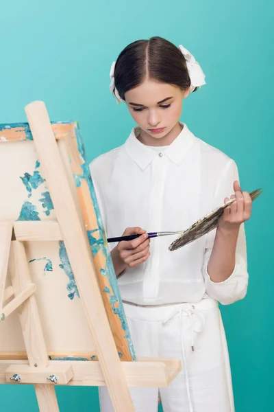 Elegant teen artist painting on easel with brush and palette, isolated on turquoise — Stock Photo