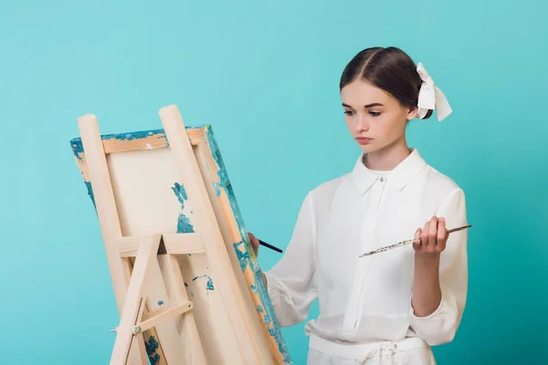 Beautiful girl painting on easel with brush and palette, isolated on turquoise — Stock Photo