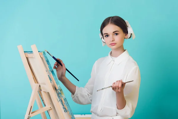 Atraente teen menina pintura no cavalete com escova e paleta, isolado no turquesa — Fotografia de Stock