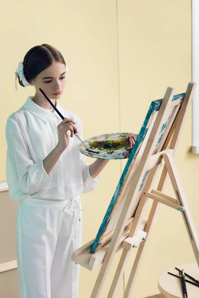 Pensive teenager painting on easel with brush and palette — Stock Photo