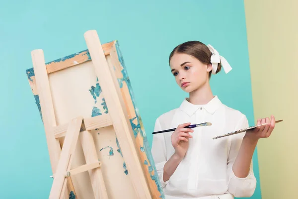 Beautiful teen artist painting on easel with brush and palette, on turquoise — Stock Photo