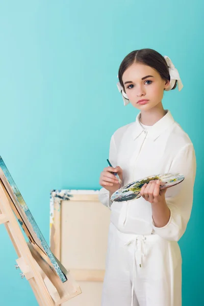Styling teen artist painting on easel with brush and palette, on turquoise — Stock Photo