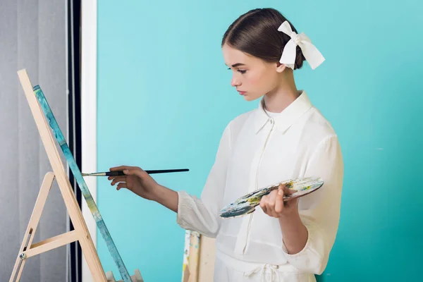 Fashionable youth artist painting on easel with brush and palette, on turquoise — Stock Photo