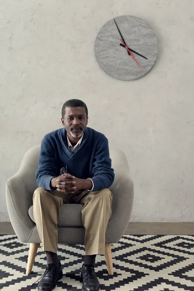 Reifer afrikanisch-amerikanischer Mann sitzt auf Sessel im Zimmer mit Uhr — Stockfoto