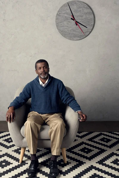 Handsome african american man sitting on armchair in room with clock — Stock Photo