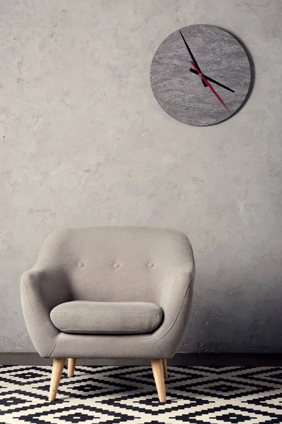 Poltrona, carpete e relógio na parede em quarto cinza simples — Fotografia de Stock