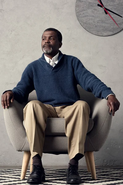 Bonito afro-americano homem sentado em poltrona com grande relógio na parede — Fotografia de Stock