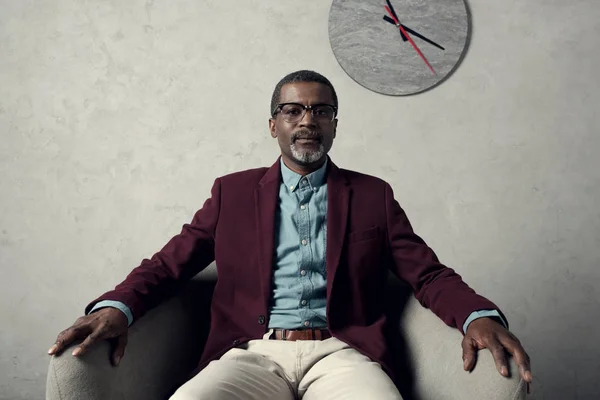 Впевнений середній вік афроамериканський чоловік сидить у кріслі з годинником на стіні — стокове фото