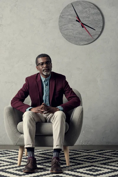Hombre afroamericano reflexivo sentado en sillón con reloj en la pared - foto de stock