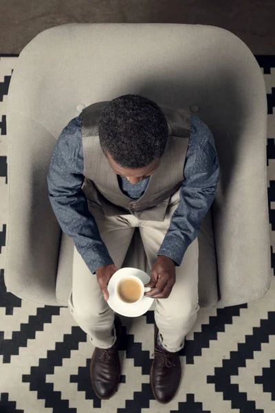 Над головой вид на стильный африканский американец, сидящий в кресле с чашкой кофе — стоковое фото