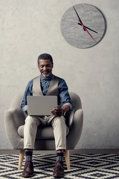 Взрослый африканский американский бизнесмен с ноутбуком сидит в кресле в офисе с часами на стене — стоковое фото