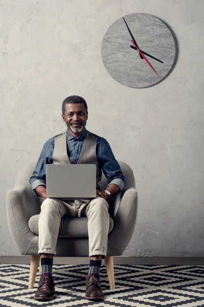 Улыбающийся африканский американский бизнесмен с помощью ноутбука в кресле в офисе с часами на стене — стоковое фото