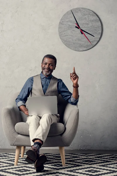 Hombre de negocios afroamericano elegante apuntando hacia arriba y usando el ordenador portátil en la oficina con el reloj en la pared - foto de stock