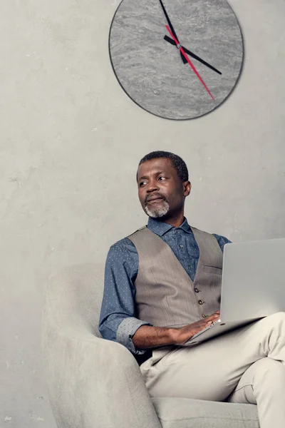 Stylish african american businessman using laptop in office with clock on wall — Stock Photo