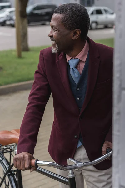 Hombre afroamericano de moda feliz en chaqueta de moda con bicicleta en la ciudad - foto de stock